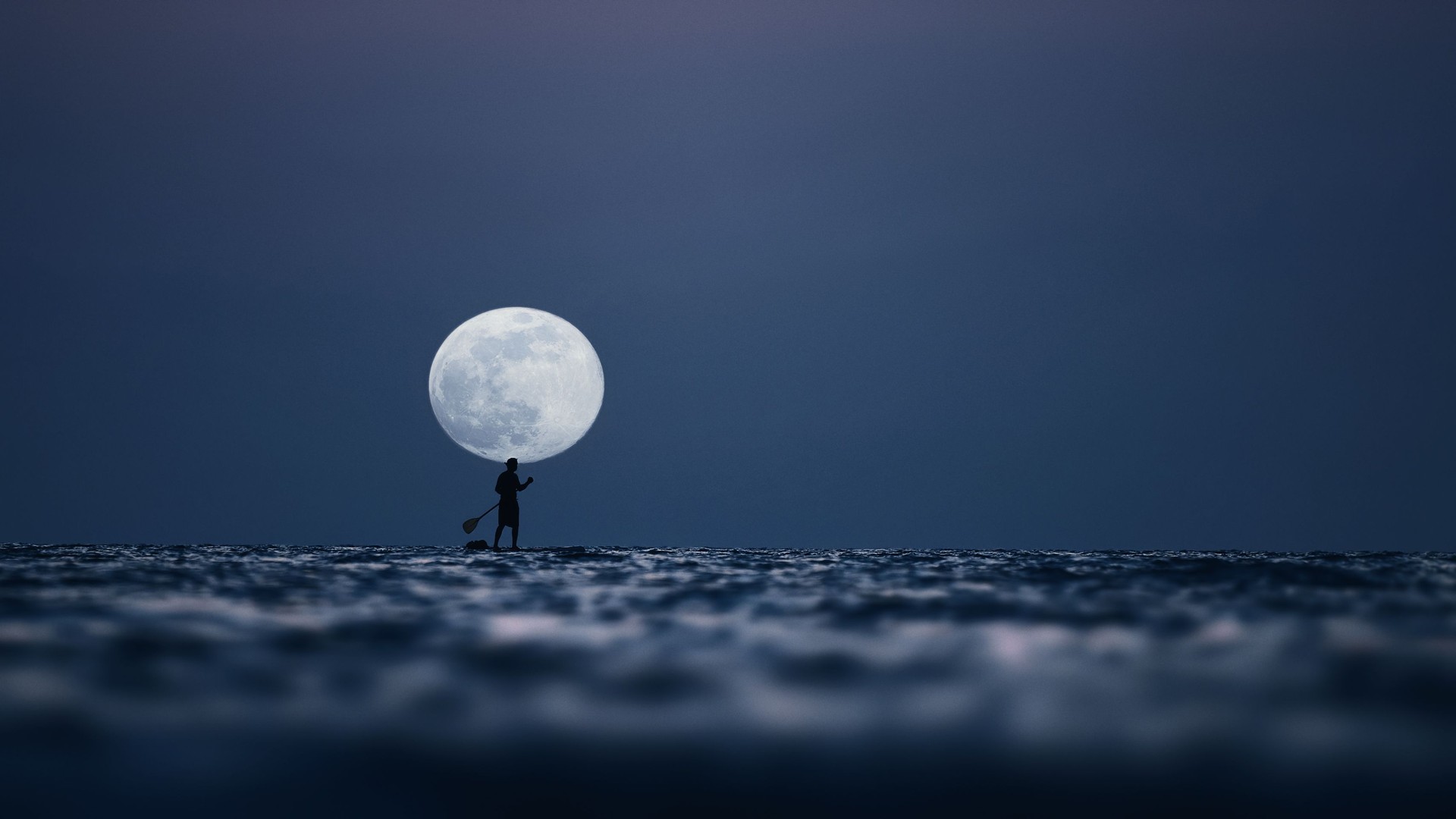 Silhouette of a man on the paddleboard against the rising moon. Paddleboard in the sea at night, side view . Young man on paddle boarding during a beautiful full moon. Paddle-boarding by open water